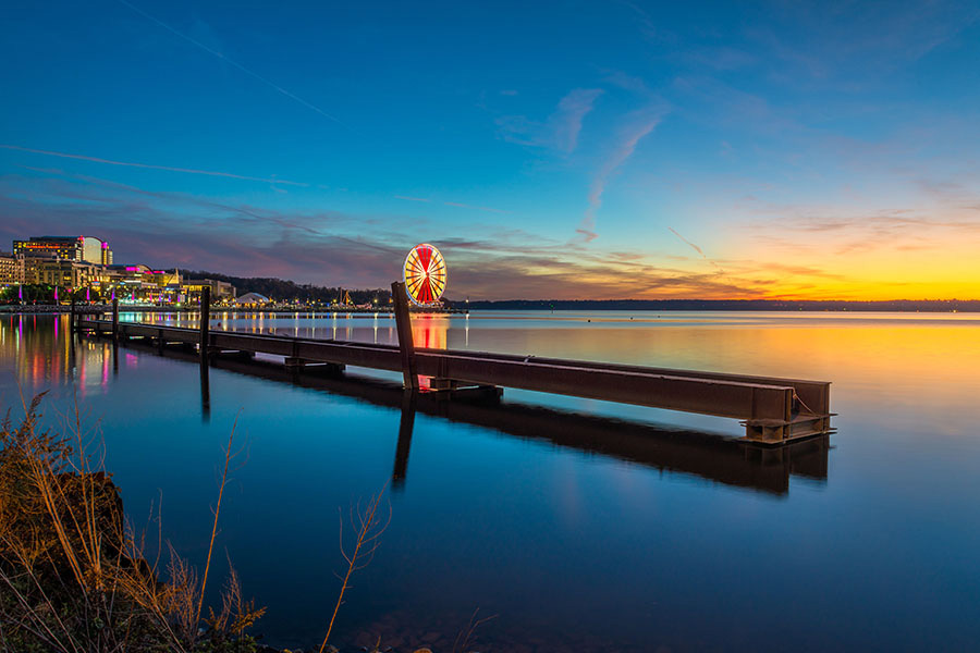 Contact - Scenic View of a Wooden Dock on a Lake with Views of the City in the Background During the Evening with a Colorful Sunset