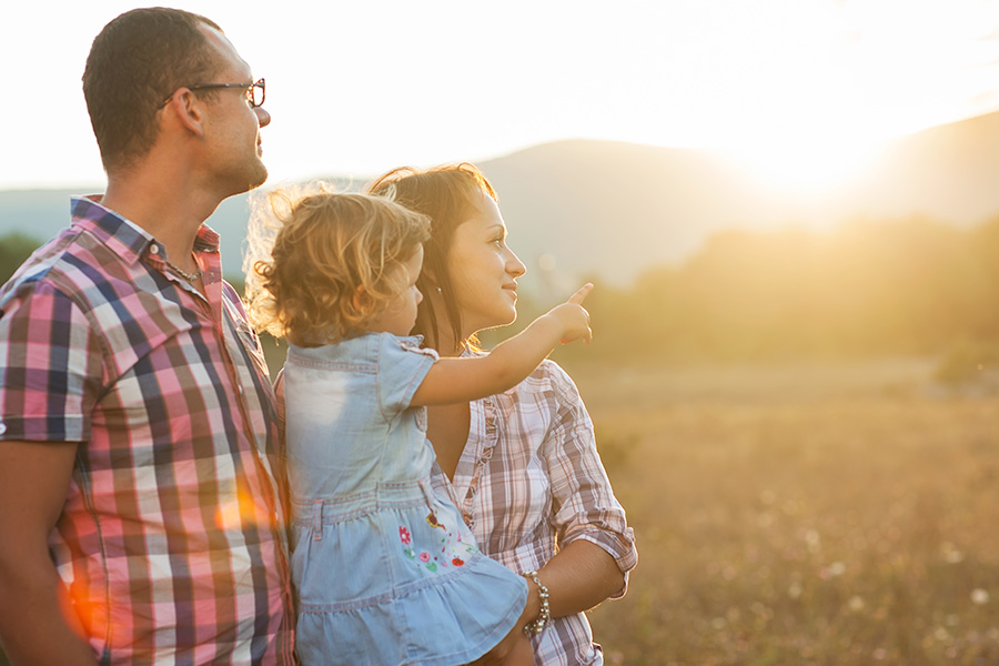 Personal Insurance - Portrait of Parents Holding Their Daughter as They Stand in a Field of Grass Looking Out at the Sunset Beyond